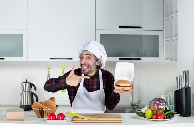 Vista frontal del chef masculino apuntando a la hamburguesa en su mano en la cocina