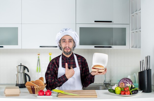 Vista frontal del chef masculino apuntando a sí mismo sosteniendo un cartel de venta y una hamburguesa en la cocina