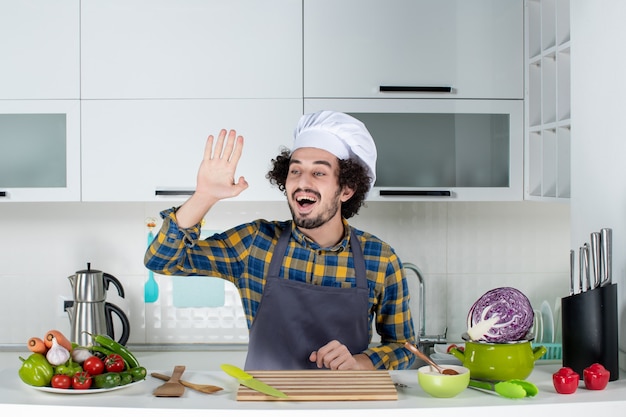 Vista frontal del chef masculino divertido y emocional con verduras frescas y cocinar con utensilios de cocina y saludar en la cocina blanca