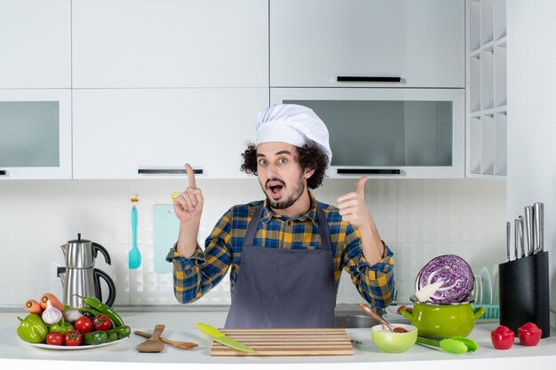 Vista frontal del chef masculino emocional con verduras frescas y cocinar con utensilios de cocina y hacer el gesto de ok en la cocina blanca