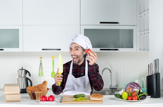 Vista frontal del chef masculino parpadeando con un cuchillo y un tomate en la cocina