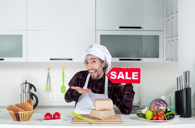 Vista frontal del chef masculino preguntado en uniforme sosteniendo cartel de venta rojo en cocina moderna