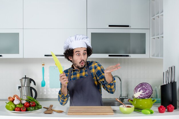 Vista frontal del chef masculino preguntándose con verduras frescas y cocinando con utensilios de cocina y sosteniendo un cuchillo en la cocina blanca