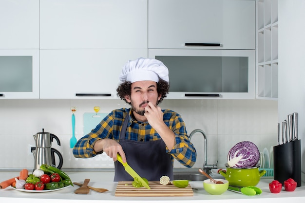 Vista frontal del chef masculino rostro amargo con verduras frescas y cocinar con utensilios de cocina y degustación de pimientos verdes en la cocina blanca