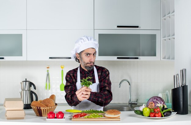 Vista frontal del chef masculino serio sosteniendo verdes en la cocina