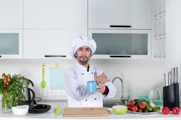 Vista frontal del chef masculino con sombrero de cocinero sosteniendo la taza de pie detrás de la mesa de la cocina