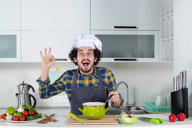 Vista frontal del chef masculino sonriente y feliz con verduras frescas degustando comida preparada y haciendo gesto de anteojos en la cocina blanca