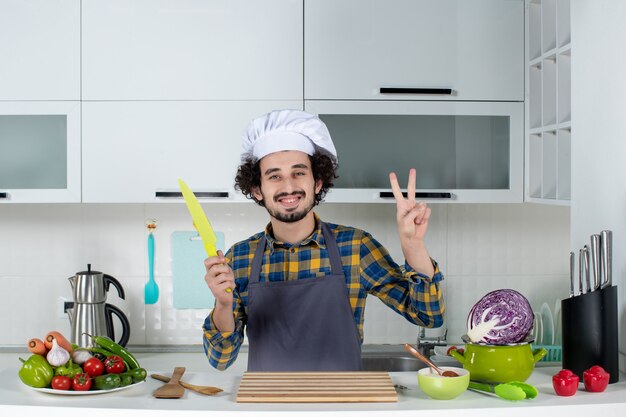 Vista frontal del chef masculino sonriente con verduras frescas y cocinar con utensilios de cocina y hacer gesto de victoria sosteniendo un cuchillo en la cocina blanca
