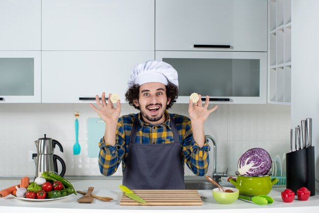 Vista frontal del chef masculino sonriente con verduras frescas y cocinar con utensilios de cocina y sosteniendo la comida en la cocina blanca