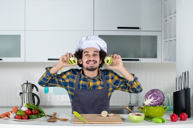 Vista frontal del chef masculino sonriente con verduras frescas y cocinar con utensilios de cocina y sosteniendo pimientos verdes en la cocina blanca