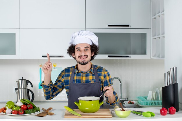Vista frontal del chef masculino sonriente con verduras frescas degustación de comida preparada y apuntando hacia arriba en la cocina blanca