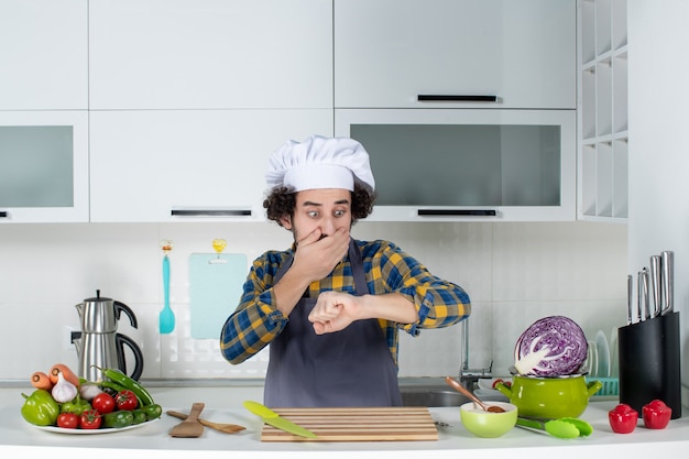 Vista frontal del chef masculino sorprendido con verduras frescas y cocinar con utensilios de cocina y controlar su tiempo en la cocina blanca