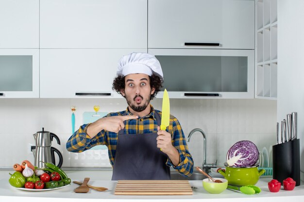 Vista frontal del chef masculino sorprendido con verduras frescas y cocinar con utensilios de cocina y un cuchillo señalador en la cocina blanca