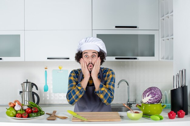 Vista frontal del chef masculino sorprendido con verduras frescas y cocinar con utensilios de cocina y posando en la cocina blanca