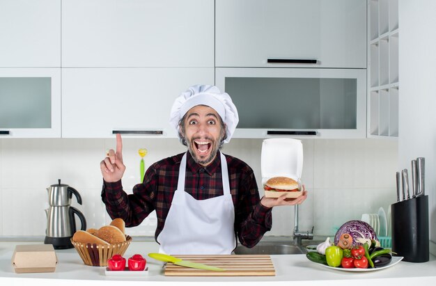 Vista frontal del chef masculino sosteniendo una hamburguesa apuntando al techo en la cocina