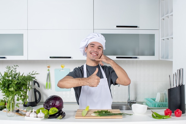 Vista frontal del chef masculino en uniforme dando pulgar hacia arriba detrás de la mesa de la cocina