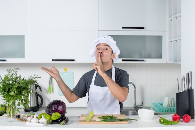 Vista frontal del chef masculino en uniforme haciendo señal de silencio detrás de la mesa de la cocina