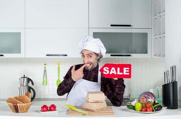 Vista frontal del chef masculino en uniforme sosteniendo cartel de venta rojo en cocina moderna