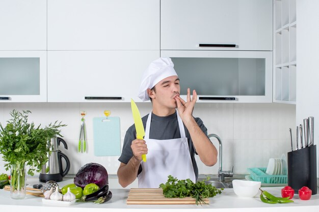 Vista frontal del chef masculino en uniforme sosteniendo un cuchillo en la cocina haciendo chef beso