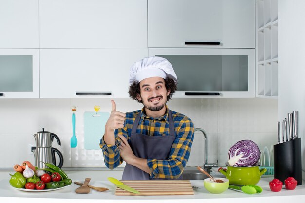 Vista frontal del chef masculino con verduras frescas y cocinar con utensilios de cocina y hacer el gesto de ok en la cocina blanca