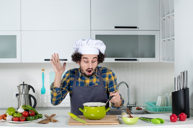 Vista frontal del chef masculino con verduras frescas y sosteniendo una cuchara en la comida y mirándola en la cocina blanca