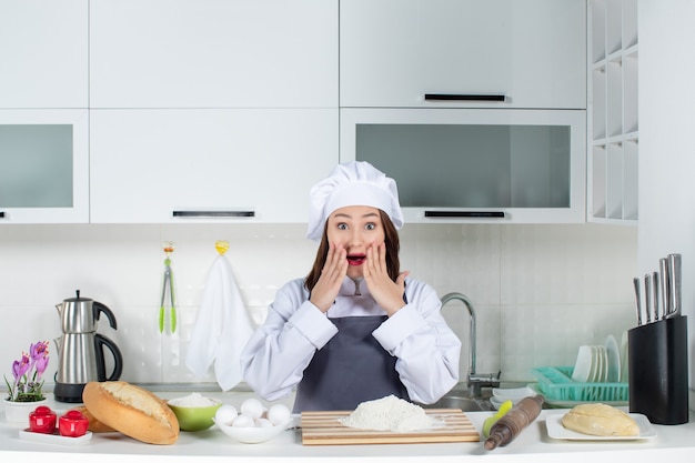 Vista frontal de la chef mujer sorprendida en uniforme de pie detrás de la mesa con verduras de pan de tabla de cortar en la cocina blanca