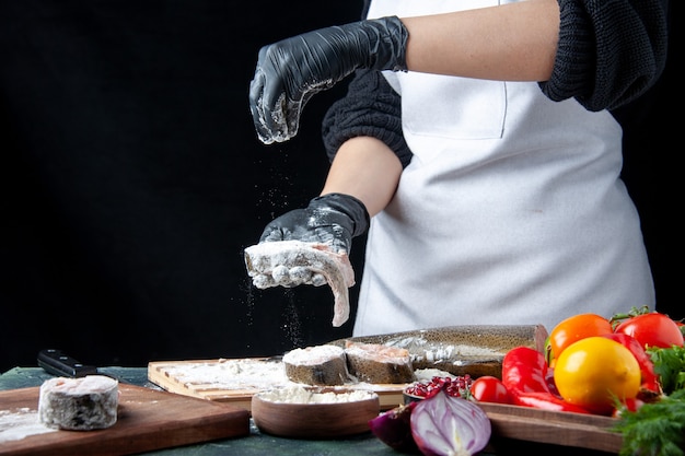 Vista frontal del chef que cubre las rodajas de pescado con verduras frescas de harina en un tazón de harina de tablero de madera en la mesa de la cocina