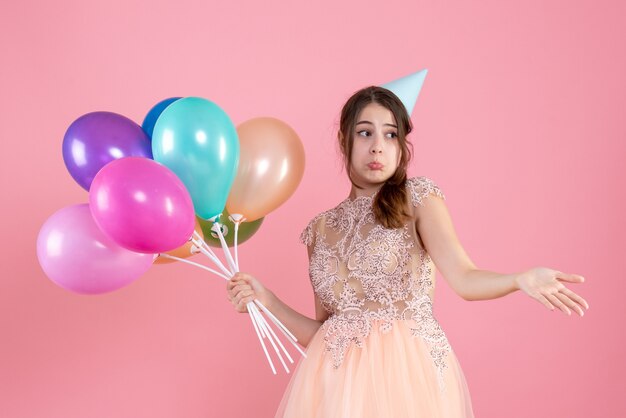 Vista frontal chica confundida con gorro de fiesta abriendo su mano sosteniendo globos