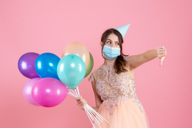 Vista frontal chica confundida con gorro de fiesta y máscara médica haciendo cartel con el pulgar hacia abajo sosteniendo globos de colores