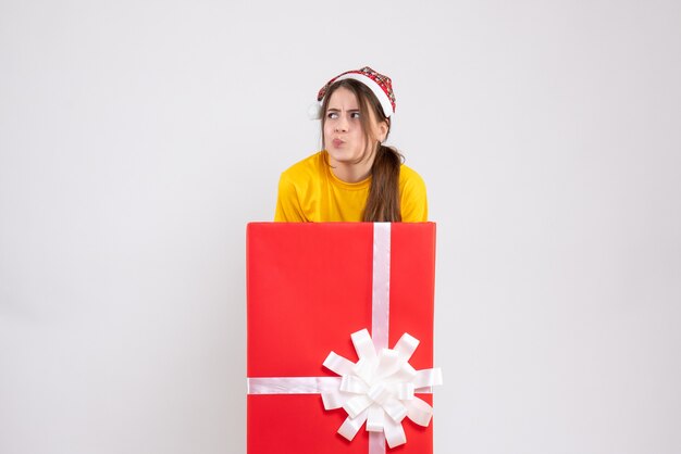 Vista frontal chica confundida con gorro de Papá Noel de pie detrás de caja de regalo de Navidad grande