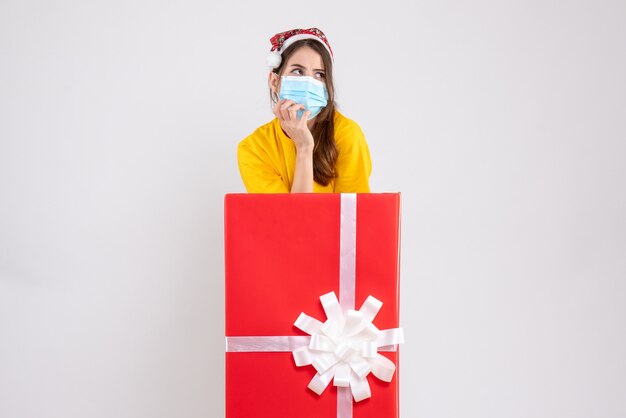 Vista frontal chica confundida con gorro de Papá Noel de pie detrás de gran regalo de Navidad