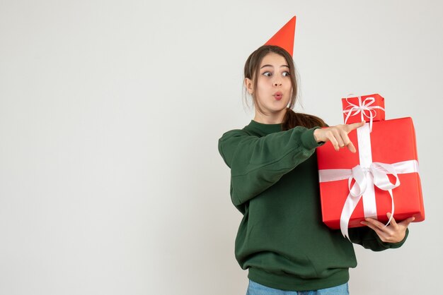 Vista frontal chica curiosa con gorro de fiesta mostrando algo sosteniendo sus regalos de Navidad