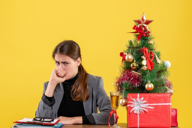 Vista frontal chica escéptica sentada en el escritorio árbol de Navidad y cóctel de regalos