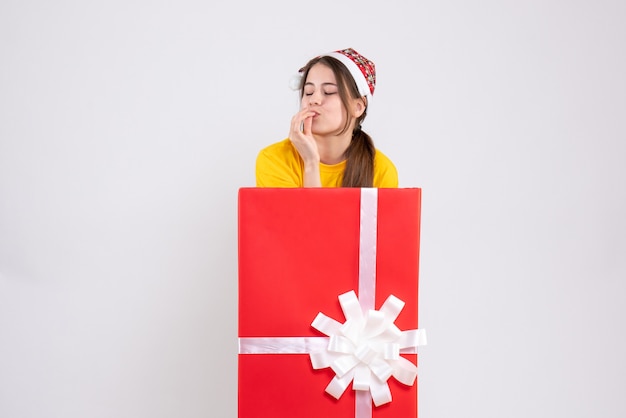 Vista frontal chica eufórica con gorro de papá noel de pie detrás de un gran regalo de navidad