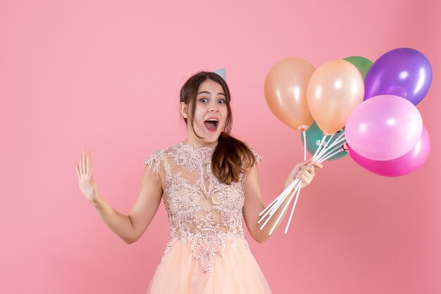 Vista frontal chica fiestera eufórica con gorro de fiesta sosteniendo globos abriendo las manos