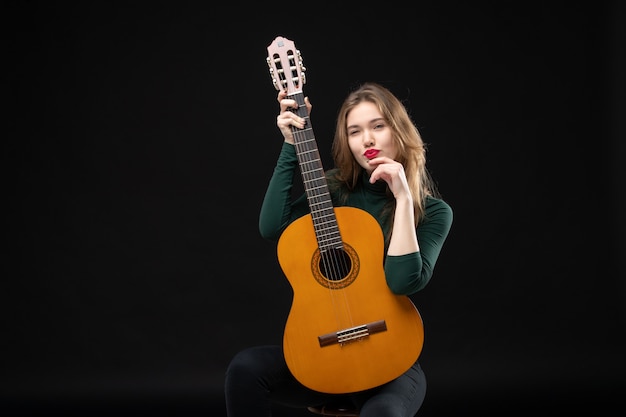 Vista frontal de la chica joven músico sosteniendo la guitarra y mirando algo cuidadosamente en la oscuridad