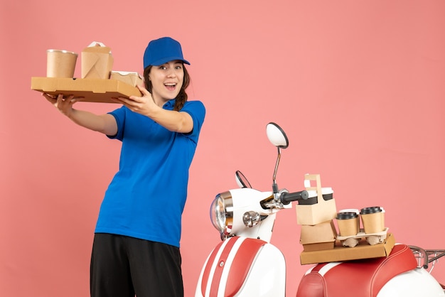 Vista frontal de la chica de mensajería de pie junto a la motocicleta mostrando café y pasteles pequeños sobre fondo de color melocotón pastel