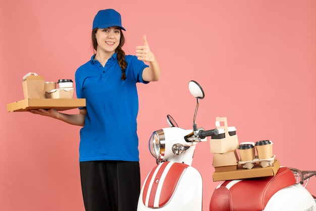 Vista frontal de la chica de mensajería de pie junto a la motocicleta sosteniendo café y pasteles pequeños haciendo gesto de ok sobre fondo de color melocotón pastel