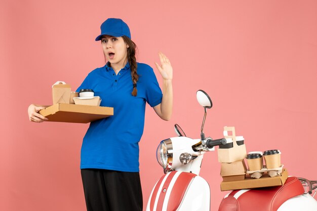 Vista frontal de la chica de mensajería sorprendida de pie junto a la motocicleta sosteniendo café y pequeños pasteles sobre fondo de color melocotón pastel