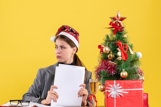 Vista frontal chica pensativa con sombrero de Navidad sentado en la mesa mirando el árbol de Navidad derecho y cóctel de regalos
