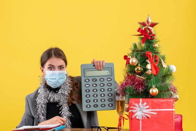 Vista frontal chica seria con máscara médica sentada en la mesa sosteniendo calculadora árbol de Navidad y cóctel de regalos