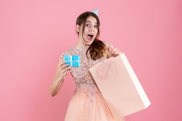 Vista frontal chica sorprendida con gorro de fiesta con bolsa y regalo