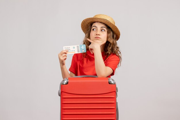 Vista frontal de la chica de vacaciones con su valija sosteniendo el boleto poniendo la mano en la barbilla
