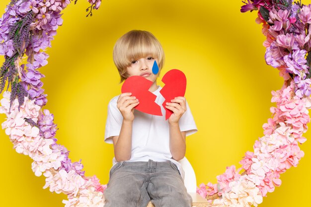 Una vista frontal chico de cabello rubio con camiseta blanca con forma de corazón sentado en la flor hecha de pie en el piso amarillo