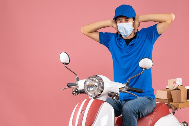 Vista frontal del chico de entrega emocional nervioso en máscara médica con sombrero sentado en scooter sobre fondo de melocotón pastel