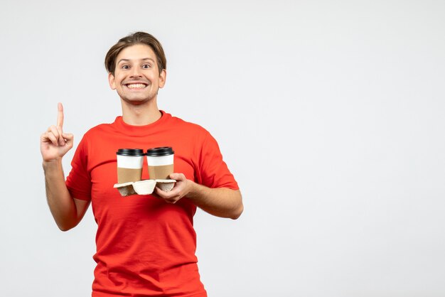 Vista frontal del chico joven feliz en blusa roja sosteniendo café en vasos de papel y apuntando hacia arriba sobre fondo blanco.