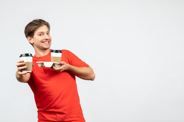Vista frontal del chico joven sonriente en blusa roja sosteniendo café en vasos de papel sobre fondo blanco.
