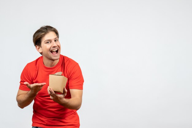 Vista frontal del chico joven sonriente y feliz en blusa roja apuntando caja pequeña sobre fondo blanco.