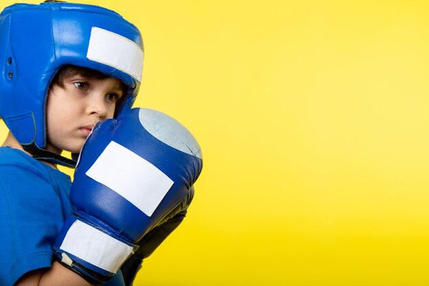 Una vista frontal chico lindo boxeo en casco azul y guantes azules en la pared amarilla