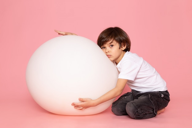 Una vista frontal chico lindo en camiseta blanca jugando con la pelota blanca en el espacio rosa